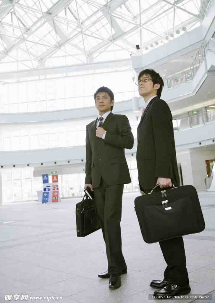 两名西装革履的男子拿着公文包,站在室内一个现代明亮的中庭,中庭有大窗户和高高的玻璃天花板,他们自信地看着前方。.